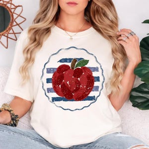 Puede incluir: Camiseta color crema con un diseño de manzana roja desgastada dentro de un círculo de rayas azules y blancas. La manzana tiene una hoja verde y un tallo marrón. La camiseta es de estilo casual y cómodo.