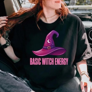 May include: Black t-shirt featuring a purple witch hat design with a crescent moon and stars. The text "BASIC WITCH ENERGY" is printed below the hat in pink. The hat has a gold buckle.