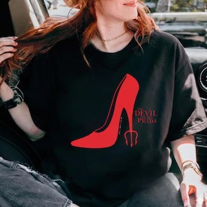 May include: Black t-shirt featuring a red high-heeled shoe graphic and the text "The Devil Wears Prada". The shoe design includes a trident. The shirt is made of a soft material.