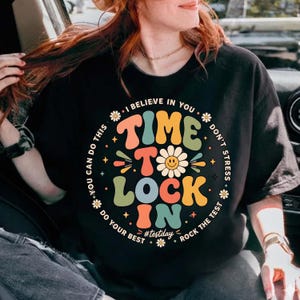 Peut inclure: T-shirt noir avec un motif graphique coloré. Le motif comprend les mots "TIME TO LOCK IN" en différentes couleurs, entourés du texte "I BELIEVE IN YOU" et "DO YOUR BEST". Une marguerite est au centre du motif.