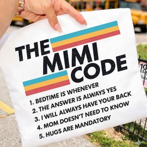 Peut inclure: Taie d'oreiller blanche avec l'inscription "THE MIMI CODE" en noir. Le coussin présente un motif de rayures horizontales colorées bleues, jaunes et rouges. Le texte en dessous énumère cinq règles, dont "L'heure du coucher est quand vous voulez".