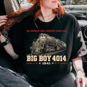May include: Black t-shirt featuring a detailed illustration of a steam locomotive, the Big Boy 4014, on a track. The shirt includes the text "AMERICAN IRON. AMERICAN LEGACY." and "BIG BOY 4014 1941" in a vintage style. The design is in shades of gray and brown.