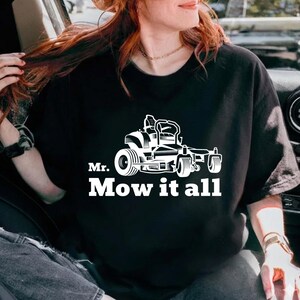 Peut inclure: T-shirt noir avec un motif blanc d'une tondeuse autoportée et le texte "Mr. Mow it all". Le motif présente une illustration détaillée de la tondeuse, avec les roues et le siège. Le texte est en gras, en police sans-serif.