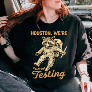 May include: Black crew neck t-shirt featuring a raccoon in a spacesuit, holding a pencil, with the text "Houston, We're Ready for Testing" in gold. The design is a humorous take on space exploration.