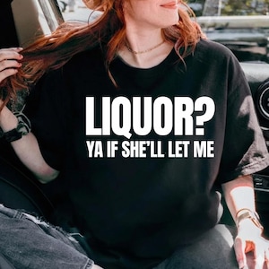 Peut inclure: T-shirt noir avec le texte blanc "LIQUOR? YA IF SHE'LL LET ME." Le t-shirt est de style col rond. La personne sur l'image porte un collier et une montre dorés.