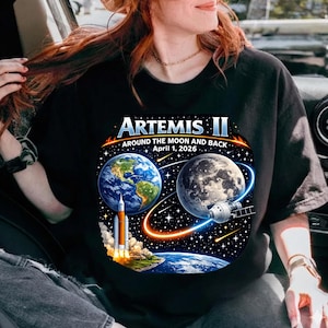 May include: Black t-shirt featuring a colorful graphic design for the Artemis II mission. The design includes Earth, the Moon, a rocket launching, and the text "Artemis II Around the Moon and Back April 1, 2026."