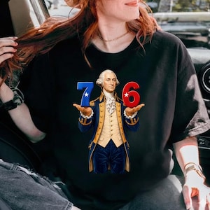Op de afbeelding: Zwart T-shirt met een afbeelding van George Washington met de cijfers "7" en "6". De "7" is blauw met een witte ster en de "6" is rood met een witte ster. Washington draagt een blauwe jas en een crèmekleurig vest.