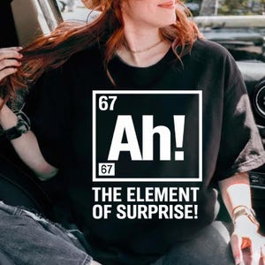 Peut inclure: T-shirt noir avec l'inscription blanche "Ah! THE ELEMENT OF SURPRISE!". Le design rappelle un élément du tableau périodique avec le chiffre 67 au-dessus et en dessous du mot "Ah!".