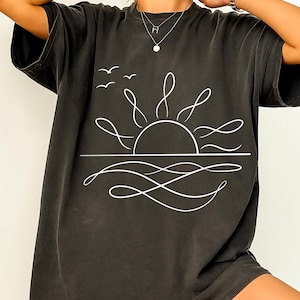 Peut inclure: T-shirt gris foncé avec un dessin au trait blanc d'un coucher de soleil sur l'océan. Le motif comprend le soleil, les vagues et trois oiseaux en vol. Le t-shirt est de style décontracté et confortable.