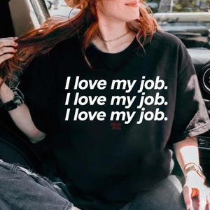 May include: Black t-shirt with the text "I love my job" repeated three times in white, with "DEVIL PRADA" in red below. The shirt is a crew neck style.