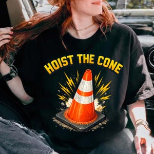 Puede incluir: Camiseta negra con el texto "HOIST THE CONE" en amarillo. El gráfico presenta un cono de tráfico naranja y blanco con un diseño de rayo amarillo. El cono está sobre una base cuadrada gris.