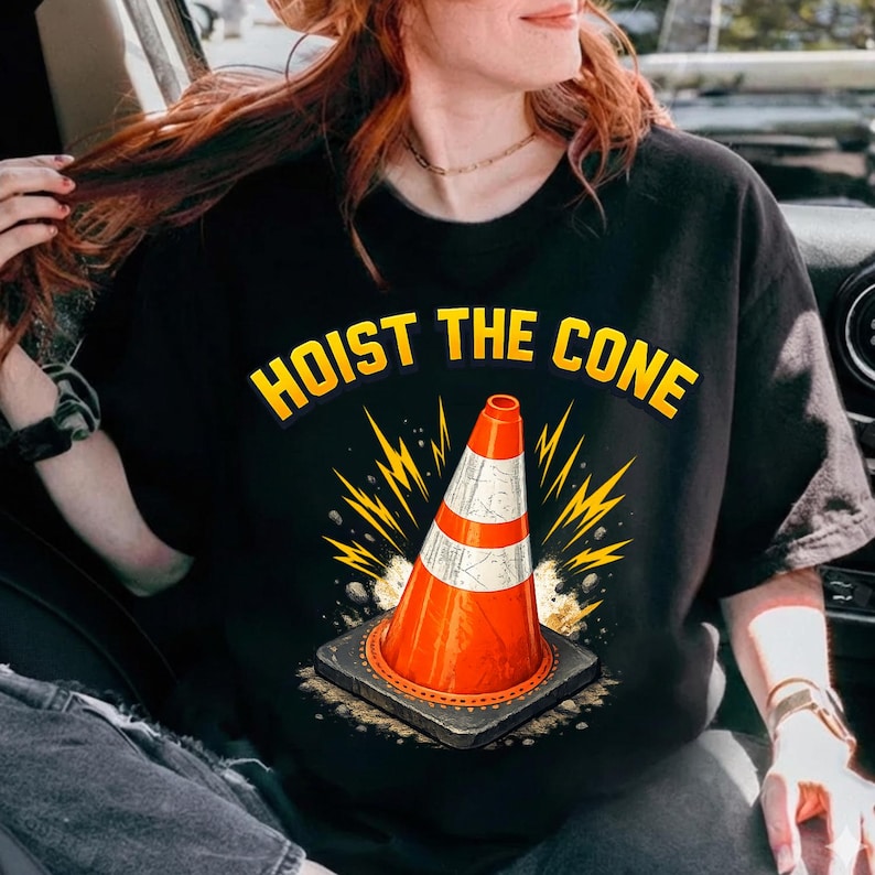 Może przedstawiać: Czarny t-shirt z ż&oacute;łtym napisem "HOIST THE CONE". Grafika przedstawia pomarańczowo-biały pachołek drogowy z ż&oacute;łtym wzorem błyskawicy. Pachołek stoi na szarej kwadratowej podstawie.