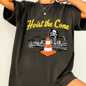 May include: Dark gray t-shirt with the text "Hoist the Cone" in yellow. The shirt features a graphic of a traffic cone with a skull and crossbones flag, set against a cityscape background.