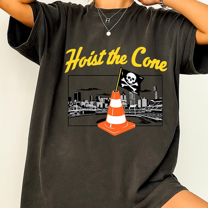 May include: Dark gray t-shirt with the text "Hoist the Cone" in yellow. The shirt features a graphic of a traffic cone with a skull and crossbones flag, set against a cityscape background.
