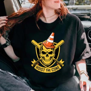 Puede incluir: Camiseta negra con una calavera amarilla que lleva un cono de tr&aacute;fico naranja y blanco. Bate de b&eacute;isbol cruzados detr&aacute;s de la calavera, con las palabras "HOIST THE CONE" debajo. El dise&ntilde;o est&aacute; sobre un escudo negro.