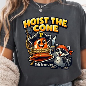 Peut inclure: T-shirt gris fonc&eacute; avec un motif de dessin anim&eacute;. Le motif comprend un c&ocirc;ne de signalisation orange souriant portant un chapeau de pirate et tenant une corde, avec un raton laveur pirate tenant un couteau. Le texte dit "HOIST THE CONE" et "This is our Jam."