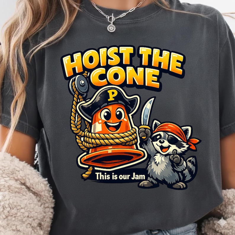 Peut inclure: T-shirt gris fonc&eacute; avec un motif de dessin anim&eacute;. Le motif comprend un c&ocirc;ne de signalisation orange souriant portant un chapeau de pirate et tenant une corde, avec un raton laveur pirate tenant un couteau. Le texte dit "HOIST THE CONE" et "This is our Jam."