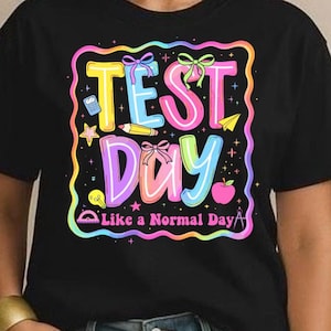 May include: Black t-shirt with a colorful graphic that says "TEST Day" in yellow, pink, and purple. The graphic includes a pencil, apple, and other school-themed elements. The text "Like a Normal Day" is below the main text.
