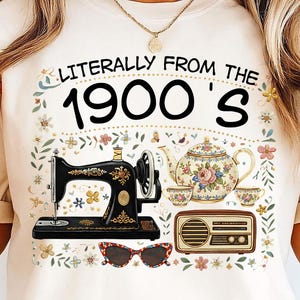 May include: Cream t-shirt featuring the text "Literally From The 1900's". The design includes a vintage sewing machine, teapot, teacups, radio, and sunglasses, with floral accents. A retro-themed garment.