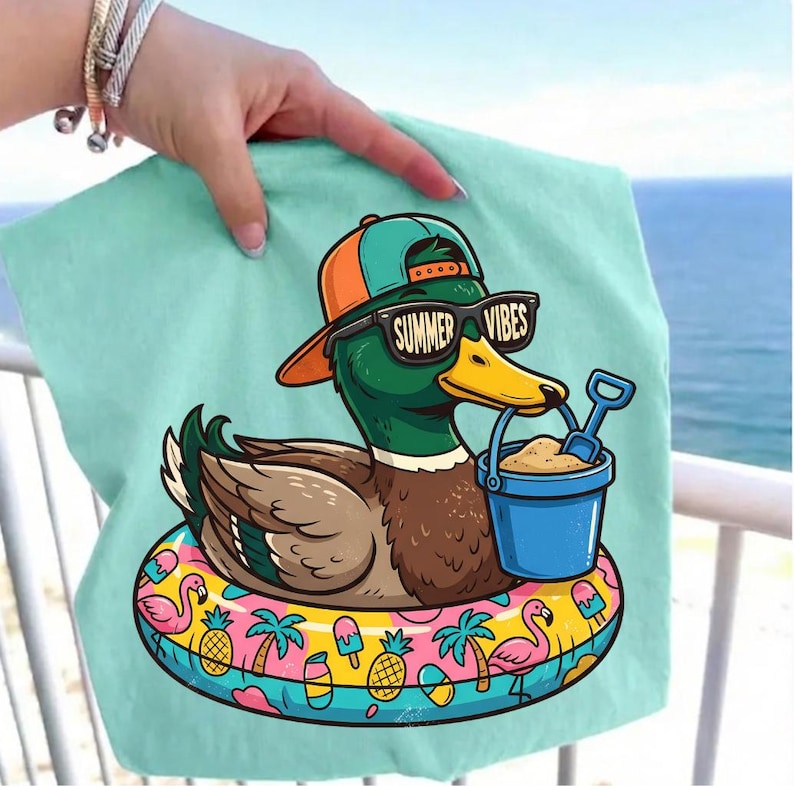 Puede incluir: Una toalla de playa de color verde azulado claro con un pato de dibujos animados que lleva gafas de sol con la inscripci&oacute;n "Summer Vibes" y una gorra de b&eacute;isbol. El pato est&aacute; en un flotador inflable colorido con pi&ntilde;as, palmeras y flamencos.