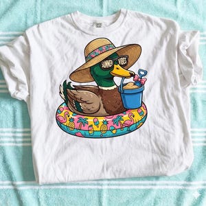 May include: White t-shirt featuring a cartoon duck wearing sunglasses with "Summer Vibes" text, a straw hat, and a beach float. The duck holds a blue bucket with sand and a shovel. The float has a colorful tropical design.