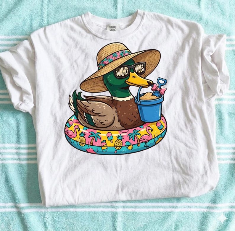 May include: White t-shirt featuring a cartoon duck wearing sunglasses with "Summer Vibes" text, a straw hat, and a beach float. The duck holds a blue bucket with sand and a shovel. The float has a colorful tropical design.