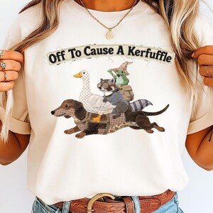 Puede incluir: Camiseta color crema con el texto "Off To Cause A Kerfuffle". El gráfico presenta un perro salchicha que lleva un ganso, un mapache y una rana. El perro es marrón, el ganso es blanco con pico amarillo, el mapache lleva gafas y la rana un sombrero.