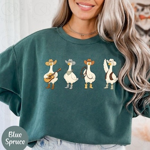 May include: Teal sweatshirt with a cartoon design of four geese in cowboy attire. Each goose wears a hat and boots, with one holding a guitar, another a lasso, and a third with a raised wing. The text "Blue Spruce" is on the bottom left of the sweatshirt.