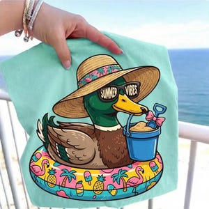 May include: A teal beach towel featuring a cartoon duck wearing sunglasses with the text "Summer Vibes" and a straw hat. The duck is floating on a colorful inflatable ring with a bucket and shovel. The background is a view of the ocean.