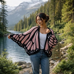 May include: A black and pink butterfly wing-shaped cardigan. The open-front sweater has a black border and a pink wing design with black detailing. The cardigan is worn with a white top and blue jeans. The background includes a lake and mountains.
