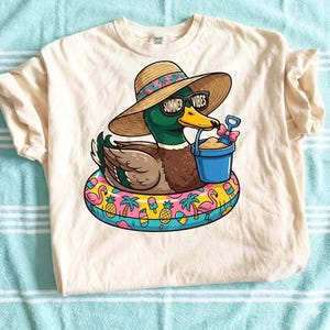 May include: Cream-colored t-shirt featuring a cartoon duck wearing sunglasses with the text "Summer Vibes" and a straw hat. The duck is floating on a colorful inflatable ring with flamingos, pineapples, and palm trees. The duck is holding a blue bucket with sand and a pink shovel.