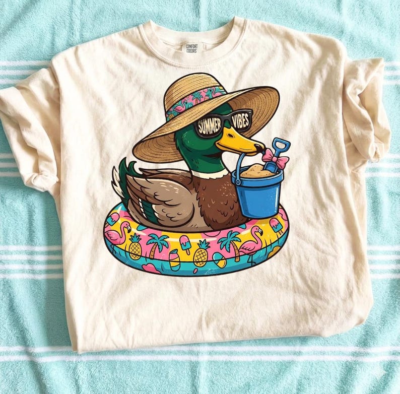 May include: Cream-colored t-shirt featuring a cartoon duck wearing sunglasses with the text "Summer Vibes" and a straw hat. The duck is floating on a colorful inflatable ring with flamingos, pineapples, and palm trees. The duck is holding a blue bucket with sand and a pink shovel.