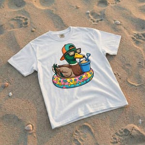 Puede incluir: Camiseta blanca con un pato de dibujos animados con gafas de sol y un sombrero, flotando en un flotador inflable. El pato sostiene un cubo azul con arena. El flotador tiene un dise&ntilde;o colorido con flamencos, pi&ntilde;as y palmeras. El texto "Summer Vibes" est&aacute; en las gafas.