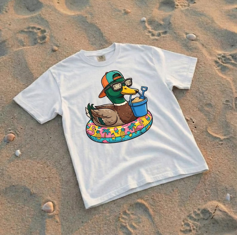 Puede incluir: Camiseta blanca con un pato de dibujos animados con gafas de sol y un sombrero, flotando en un flotador inflable. El pato sostiene un cubo azul con arena. El flotador tiene un dise&ntilde;o colorido con flamencos, pi&ntilde;as y palmeras. El texto "Summer Vibes" est&aacute; en las gafas.