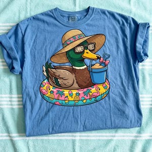 May include: A light blue t-shirt featuring a cartoon duck wearing sunglasses and a straw hat with the text "Summer Vibes." The duck is floating on a colorful inflatable ring with tropical designs, holding a bucket and shovel.