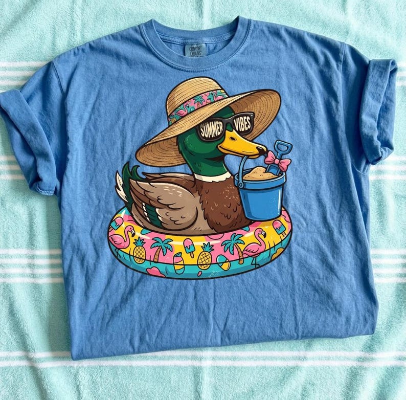 May include: A light blue t-shirt featuring a cartoon duck wearing sunglasses and a straw hat with the text "Summer Vibes." The duck is floating on a colorful inflatable ring with tropical designs, holding a bucket and shovel.