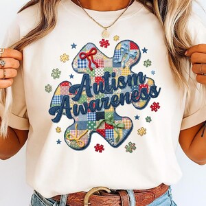 May include: Cream-coloured t-shirt featuring a colourful patchwork puzzle piece with the words "Autism Awareness" in blue script. The design includes small puzzle pieces and stars. The shirt has a round neckline and short sleeves.