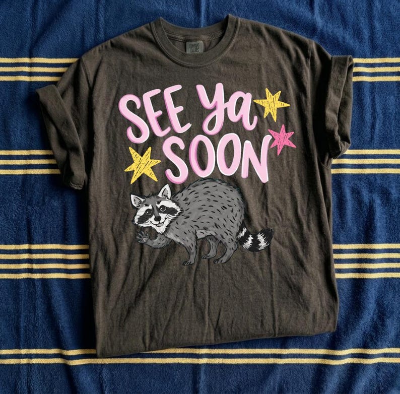 Może przedstawiać: Ciemnobrązowa koszulka z napisem "SEE ya SOON" w kolorze r&oacute;żowym, ż&oacute;łtymi i r&oacute;żowymi gwiazdami oraz ilustracją z kresk&oacute;wkowym szopem. Koszulka jest złożona na niebiesko-złotej powierzchni w paski.