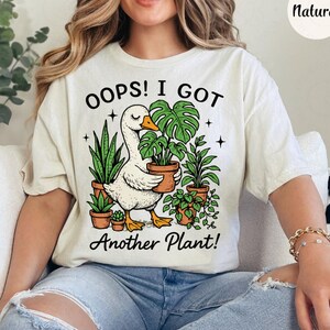 Puede incluir: Camiseta color crema con el texto "Oops! I Got Another Plant!" y un ganso de dibujos animados sosteniendo una planta en maceta. El diseño incluye varias plantas en maceta y estrellas, creando una estética caprichosa con temática de plantas.