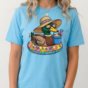 May include: Light blue t-shirt featuring a cartoon duck wearing sunglasses with the text "Summer Vibes" and a straw hat. The duck is floating on a colorful inflatable ring with a bucket of sand. The shirt is a summer-themed design.