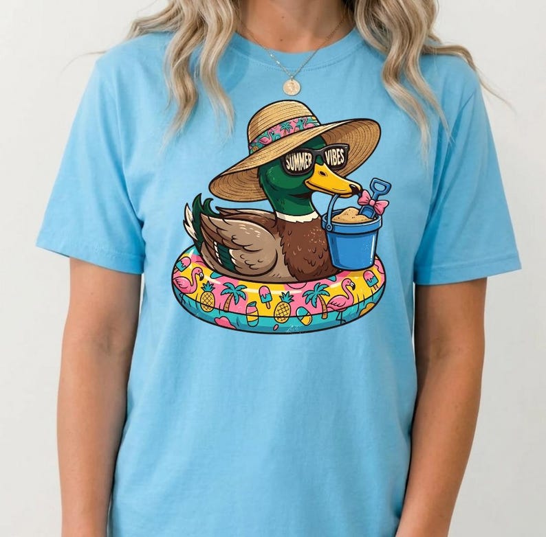 May include: Light blue t-shirt featuring a cartoon duck wearing sunglasses with the text "Summer Vibes" and a straw hat. The duck is floating on a colorful inflatable ring with a bucket of sand. The shirt is a summer-themed design.