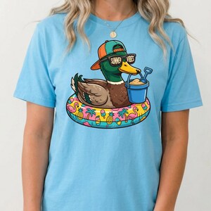 Puede incluir: Camiseta azul claro con un pato de dibujos animados con gafas de sol y gorra, con el texto "Summer Vibes". El pato est&aacute; en un flotador inflable con dise&ntilde;os tropicales, sosteniendo un cubo y una pala.