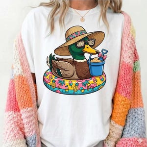 May include: White t-shirt featuring a cartoon duck wearing sunglasses that read "Summer Vibes" and a straw hat. The duck is floating on a colorful inflatable ring with beach-themed graphics. The duck holds a blue bucket with sand and a small shovel.