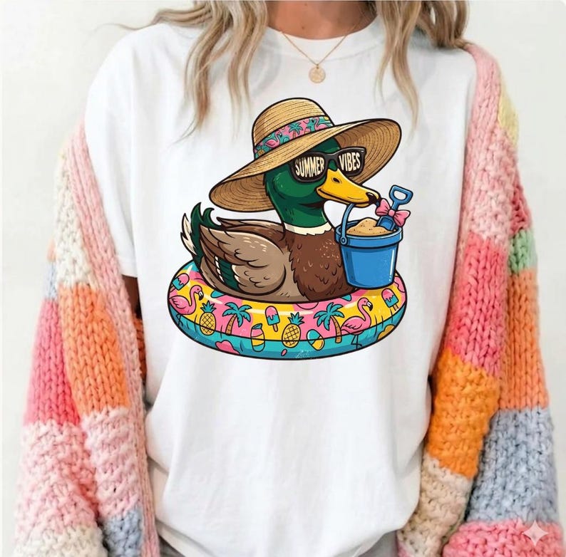 May include: White t-shirt featuring a cartoon duck wearing sunglasses that read "Summer Vibes" and a straw hat. The duck is floating on a colorful inflatable ring with beach-themed graphics. The duck holds a blue bucket with sand and a small shovel.