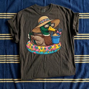 May include: Dark gray t-shirt featuring a cartoon duck wearing sunglasses and a sun hat with the text "Summer Vibes." The duck is floating on a colorful inflatable ring with tropical designs and holding a blue bucket with sand.