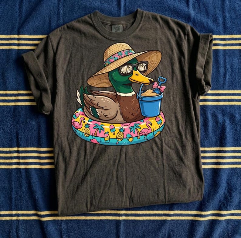 May include: Dark gray t-shirt featuring a cartoon duck wearing sunglasses and a sun hat with the text "Summer Vibes." The duck is floating on a colorful inflatable ring with tropical designs and holding a blue bucket with sand.