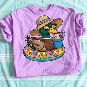 May include: A lilac-colored t-shirt featuring a cartoon duck wearing sunglasses that read "Summer Vibes" and a straw hat. The duck is floating on a colorful inflatable ring with a bucket and shovel. The shirt is made of a soft material.