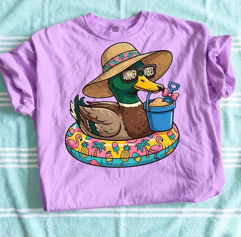 May include: A lilac-colored t-shirt featuring a cartoon duck wearing sunglasses that read "Summer Vibes" and a straw hat. The duck is floating on a colorful inflatable ring with a bucket and shovel. The shirt is made of a soft material.
