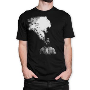 May include: Black t-shirt featuring a black and white graphic of a person smoking, with smoke forming a cloud and birds. The person is wearing a button-down shirt. The design is centered on the chest.