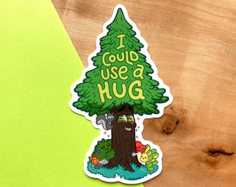 Könnte beinhalten: Ein grüner Cartoon-Baum mit einem lächelnden Gesicht und dem Text "I could use a hug" am Stamm. Der Baum ist von kleinen Cartoon-Tieren und Pflanzen umgeben.